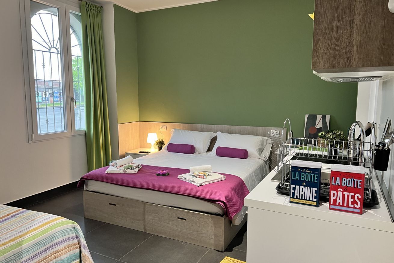 Een hotelkamer met een bed, een nachtkastje, een raam met gordijnen en een kitchenette met een afwasschaal en een bordje.