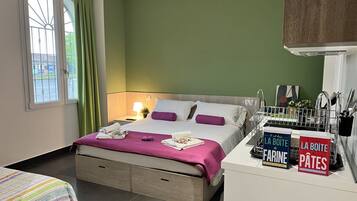 Een hotelkamer met een bed, een nachtkastje, een raam met gordijnen en een kitchenette met een afwasschaal en een bordje.