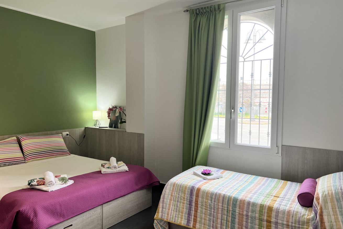 Een hotelkamer met twee bedden, een raam met groene gordijnen, een nachtkastje met een lamp en uitzicht op buiten.