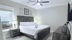 Un dormitorio con cama, mesitas de noche, ventilador de techo, ventana y televisor sobre un mueble.