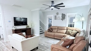 Una sala de estar con un sofá color beige, un sillón blanco y un sofá marrón. Hay un televisor de pantalla plana sobre un mueble de madera, un ventilador de techo y una escalera que sube a otro nivel.