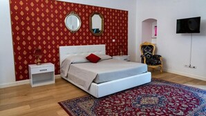 A bedroom with a bed, two bedside tables, a TV, a mirror, a patterned rug, and a chair. - Palazzo delle Puglie B&B (Barletta)