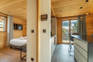 A wooden cabin interior with a bed, a desk, a chair, and a view of the outdoors.