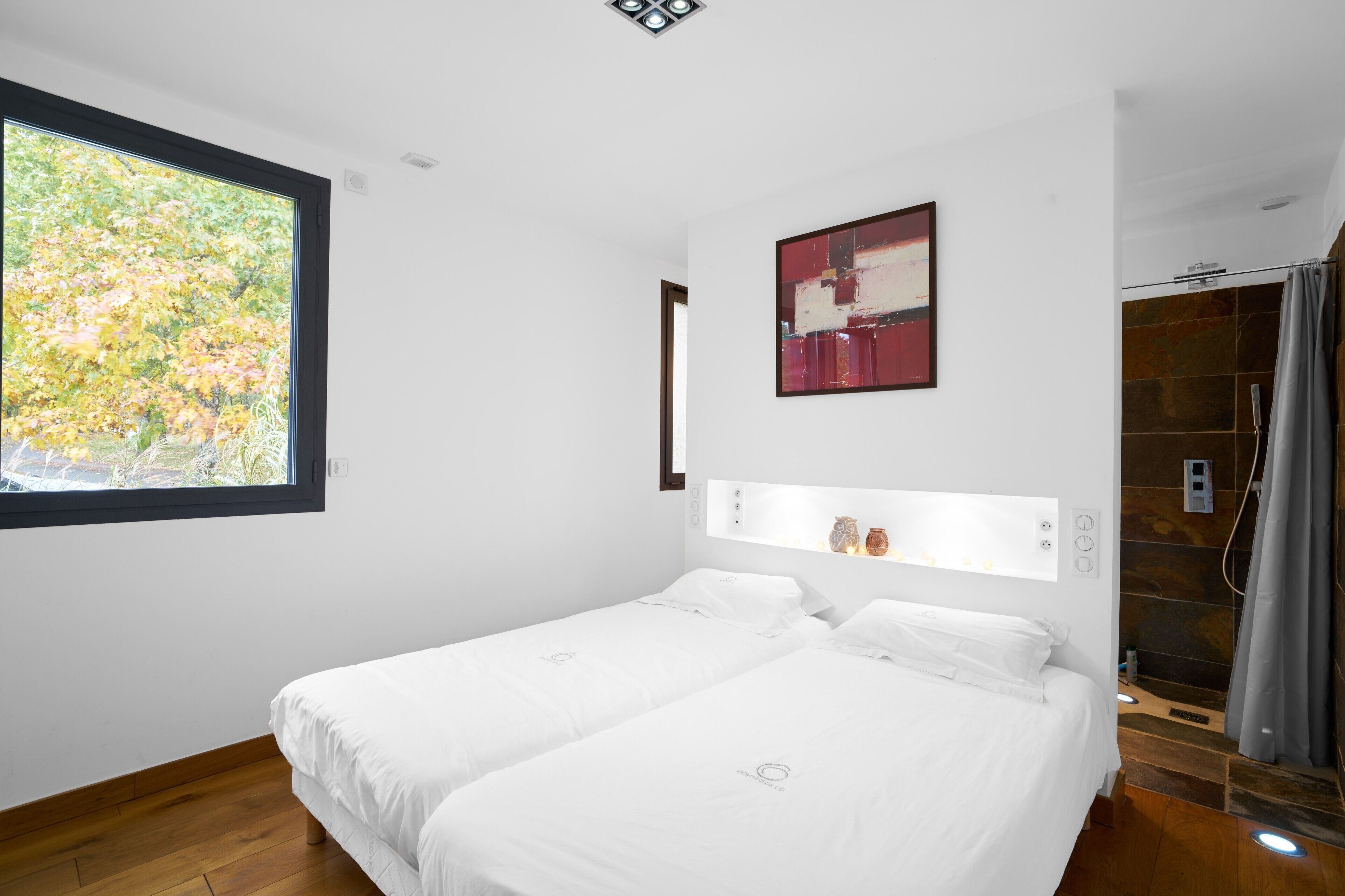 Une chambre d’hôtel moderne avec deux lits, une grande fenêtre, un espace douche et une étagère fixée au mur, agrémentée d’objets décoratifs.
