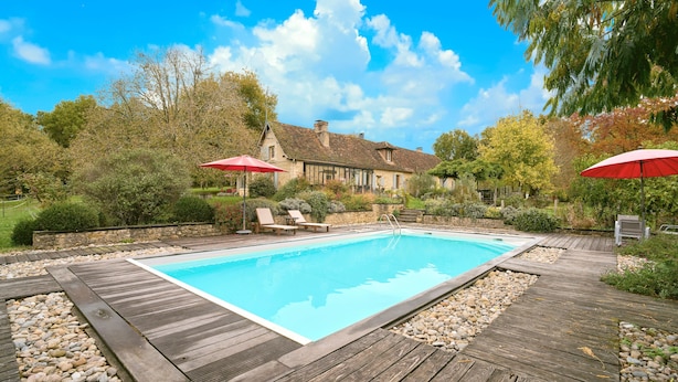 A swimming pool with wooden decking, surrounded by a stone border, and two red umbrellas. - Loft Ôvebas at Domaine De L'ô (Eyraud-Crempse-Maurens)