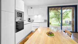 A modern kitchen with white cabinetry, a wooden dining table, and a view of the outdoors through a glass door. - Loft Ôvebas at Domaine De L'ô (Eyraud-Crempse-Maurens)