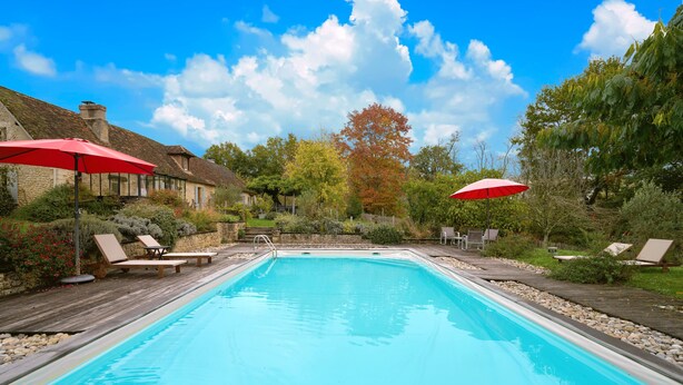 Une piscine rectangulaire entourée d’une terrasse en bois, avec deux parasols rouges et des chaises longues.