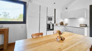 Une cuisine moderne avec des meubles blancs, une table à manger en bois et une vue dégagée sur l’extérieur grâce à une grande fenêtre.