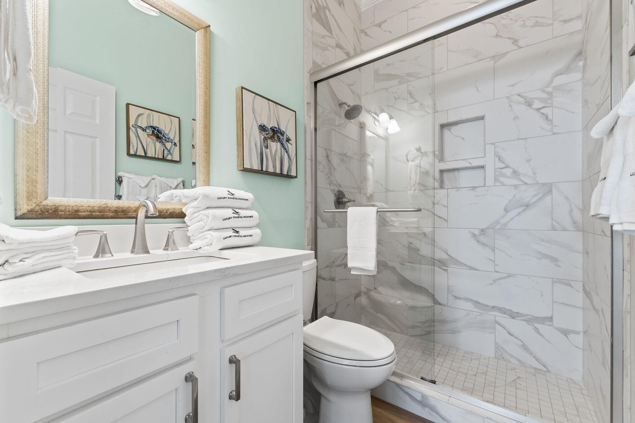 A bathroom with a white vanity, a toilet, a glass shower enclosure, and two framed artworks on the wall.