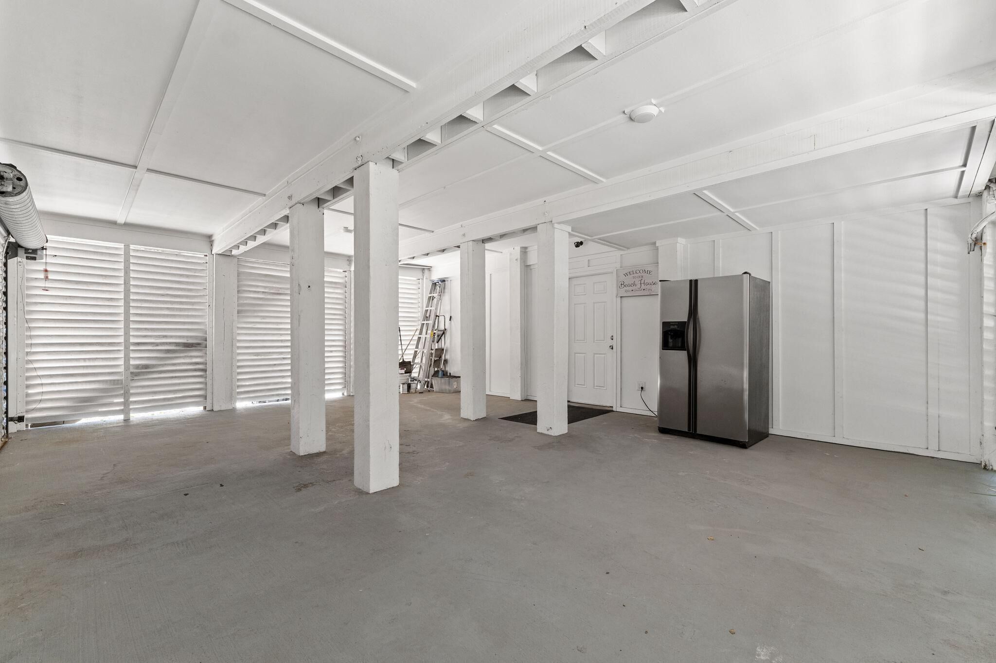 A spacious, industrial-style room with white walls and ceiling, featuring a stainless steel refrigerator and a ladder leaning against the wall.