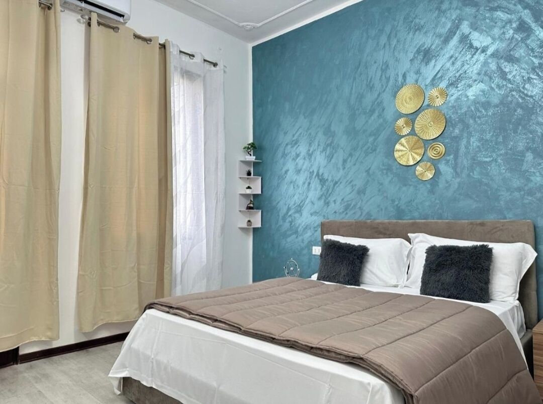 A bedroom with a bed, beige curtains, a teal accent wall, and a small shelf with decorative items.