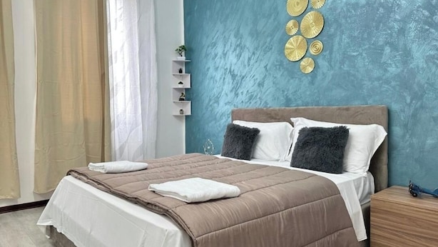 A bedroom with a bed, a nightstand, a wall with decorative plates, and a window with curtains. - Venetian Nexus (Mestre)