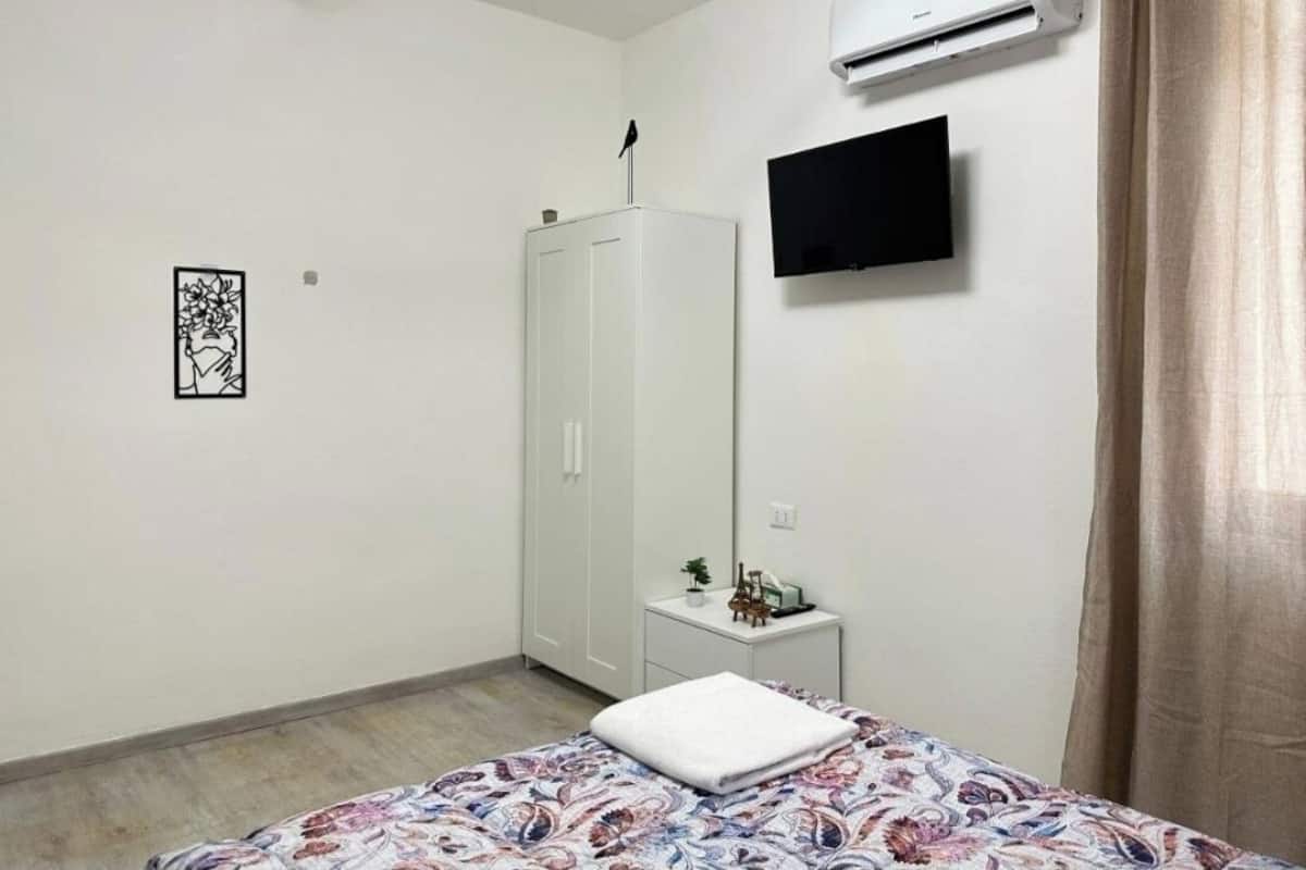 A bedroom with a bed, a wardrobe, a small bedside table with a plant, a TV mounted on the wall, and an air conditioning unit.