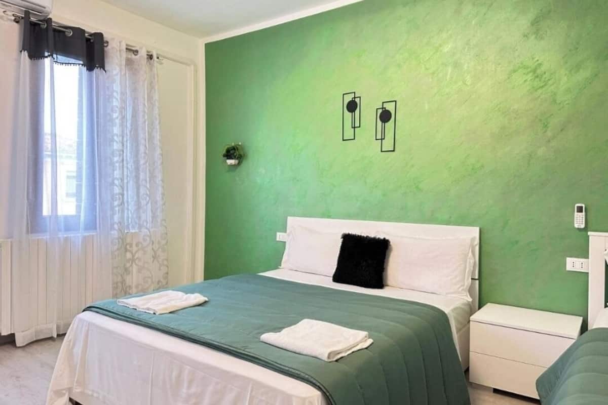 A bedroom with a green wall, a bed with white and green bedding, a black pillow, a white bedside table, and a white nightstand.