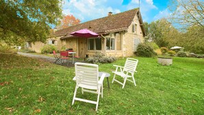 Une maison en pierre avec un toit en tuiles, une terrasse avec deux chaises blanches et une table, et un parasol violet.