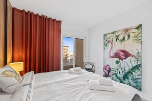 A hotel room with a bed, a painting of a flamingo, a window with curtains, and a view of buildings outside.