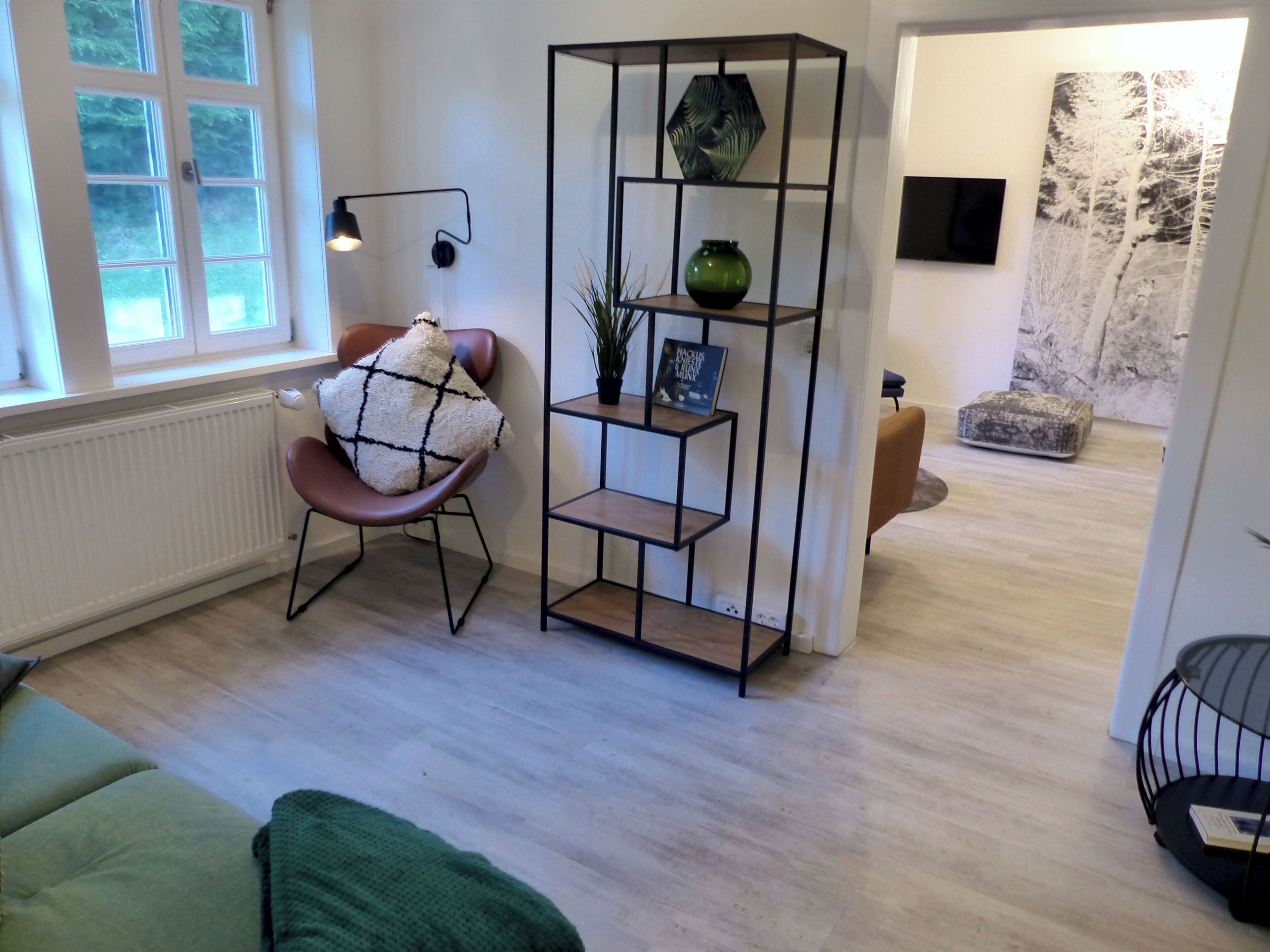 A modern living room with a wooden floor, a green sofa, a brown chair with a patterned cushion, a black metal shelving unit with decorative items, and a window with blinds.