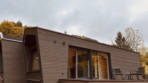 Un edificio moderno de madera con un amplio ventanal corredera, un pequeño espacio de estar al aire libre y una cocina visible.