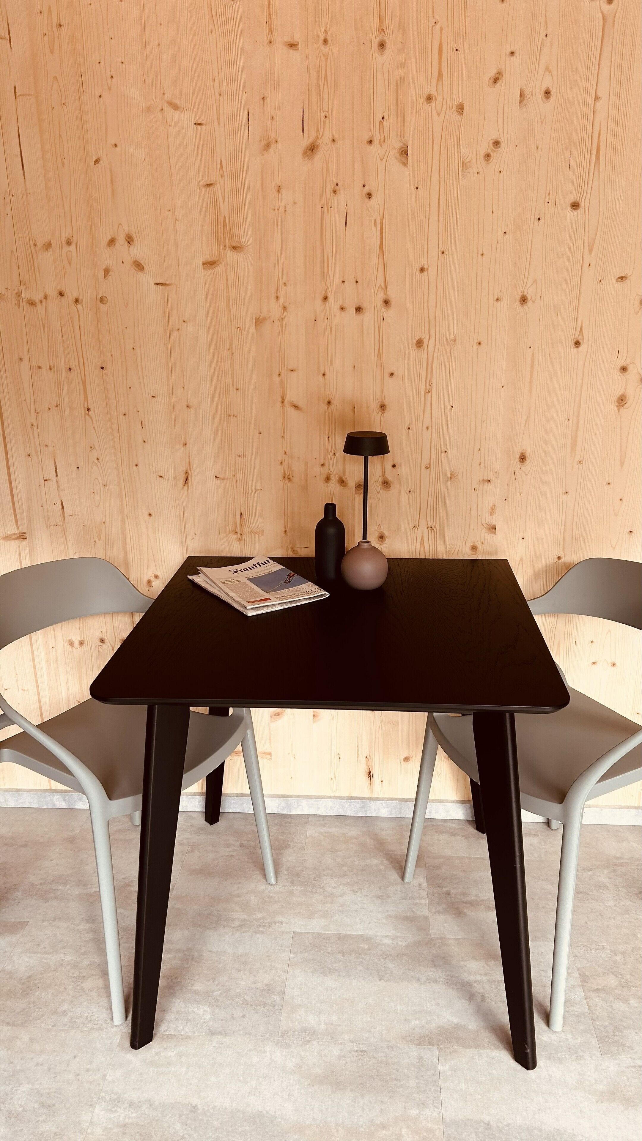 A table with a black top, two grey chairs, a black bottle, and a vase on a wooden wall background.