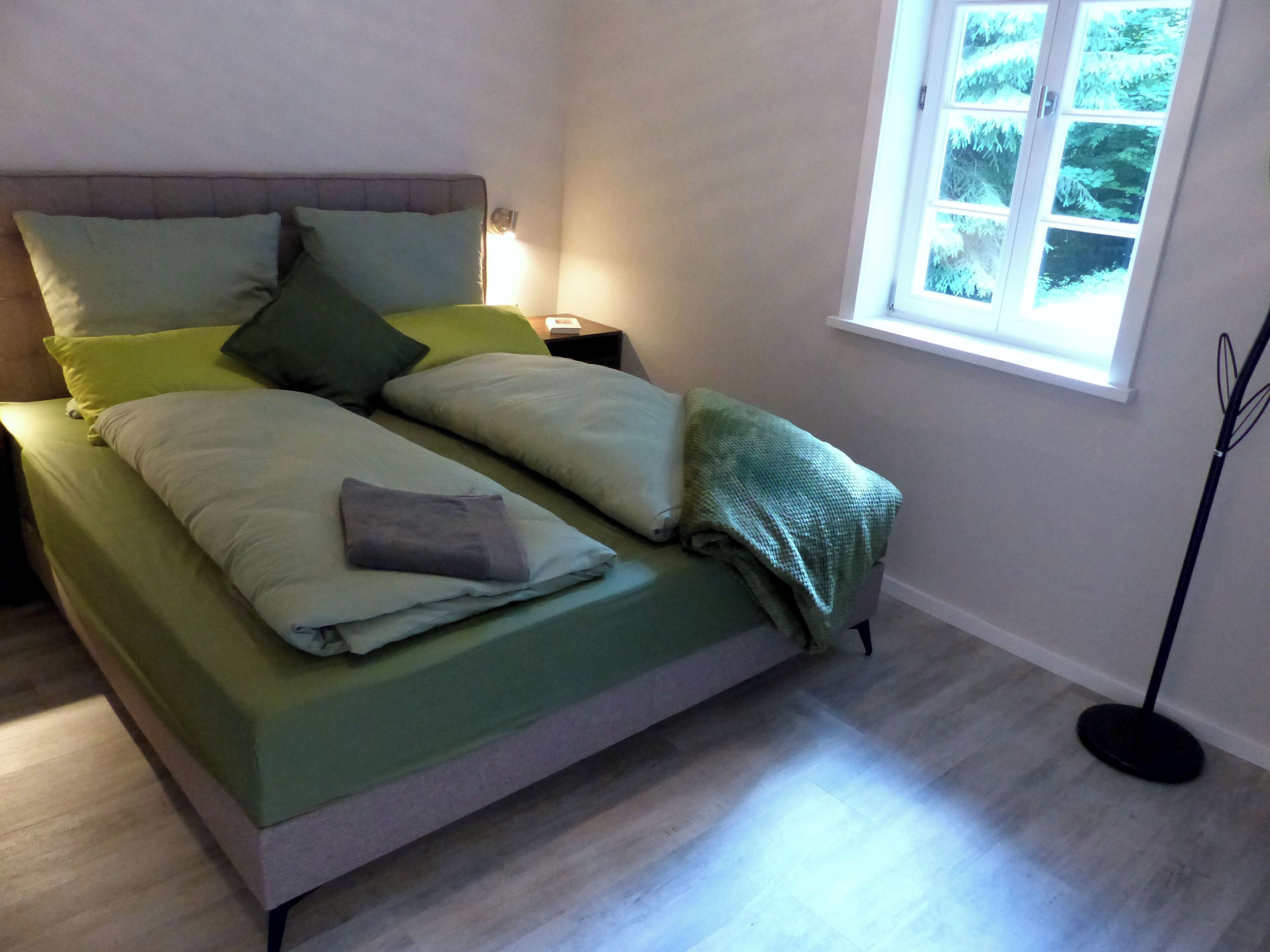 Un letto rifatto con cura, con biancheria verde e numerosi cuscini, un comodino con una lampada e una finestra vista alberi.