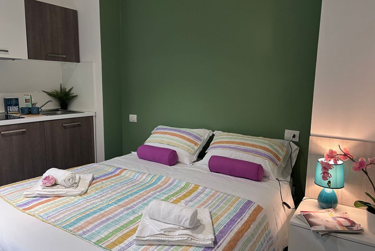 Habitación de hotel con cama, almohadas, mesita de noche, lámpara y una planta pequeña.