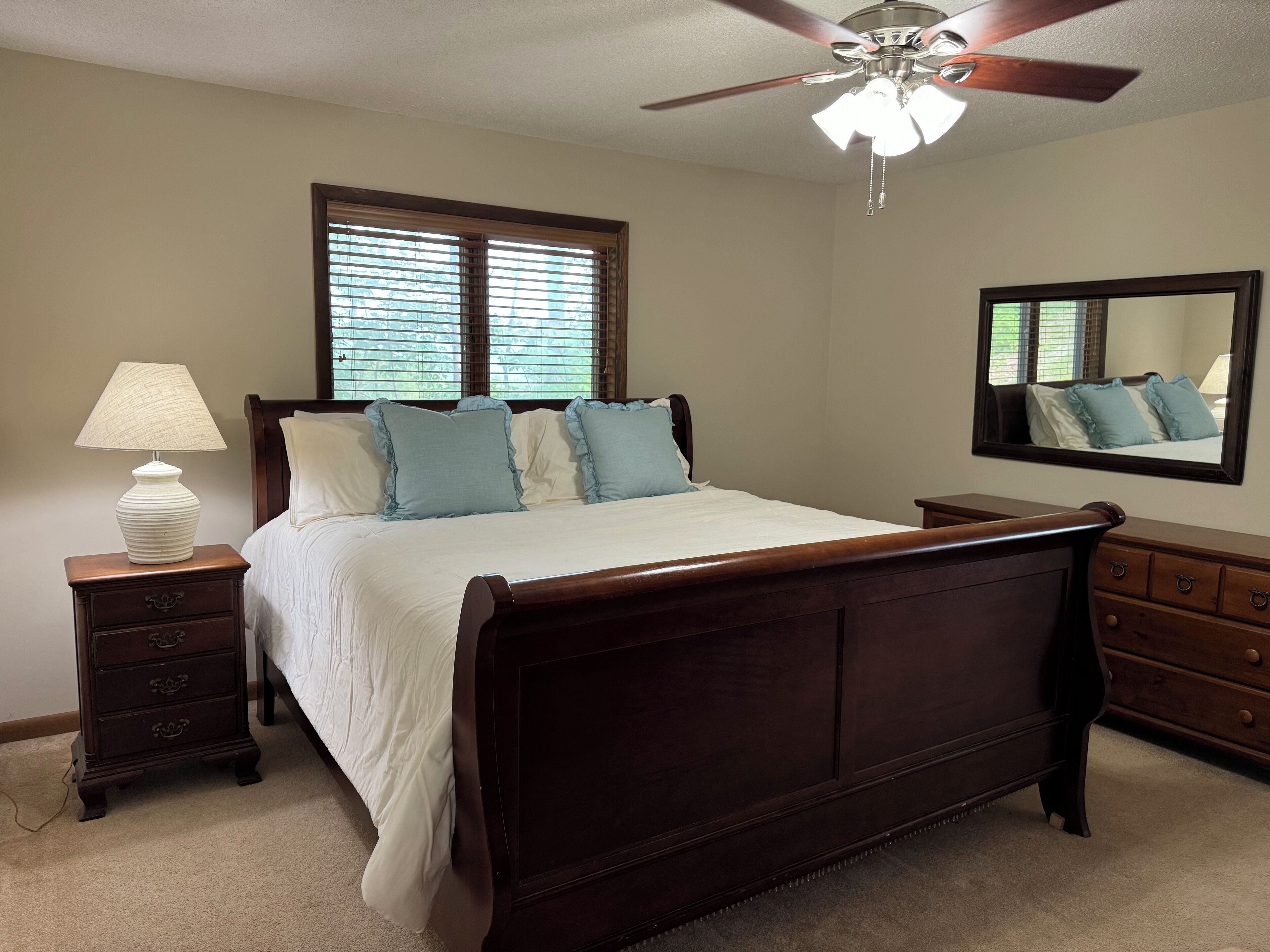 Une chambre avec un lit en bois, une table de chevet avec une lampe, une commode, un ventilateur de plafond et une fenêtre avec des stores.
