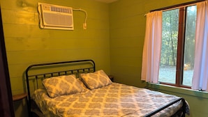 A single bed with a patterned bedspread, a wall-mounted air conditioner, and a window with curtains.