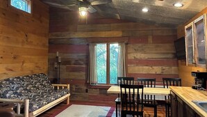 A cozy cabin interior with a wooden log wall, a sofa, a dining table with chairs, and a ceiling fan.