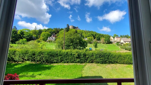 Une vue prise de l’intérieur d’une pièce, donnant sur un paysage verdoyant avec un château perché sur une colline, des maisons et une haie.