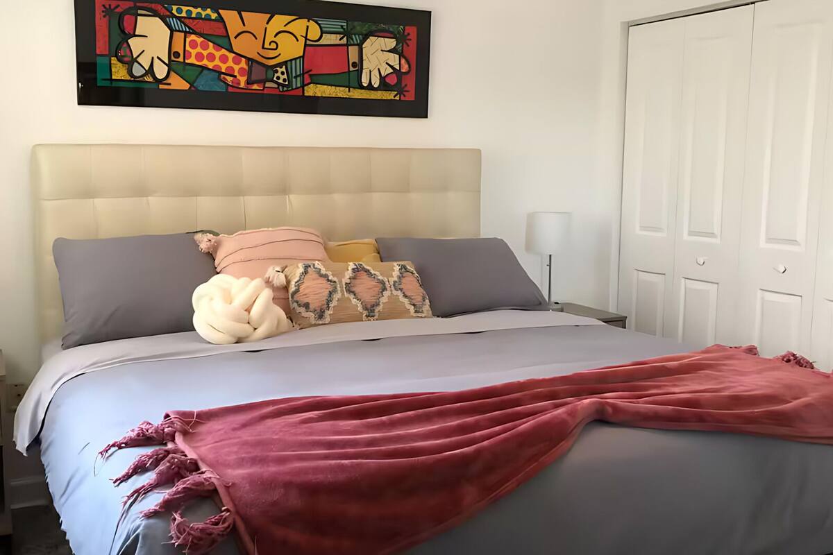 Une chambre à coucher avec un lit, des oreillers, une couverture, une table de chevet, une lampe et un tableau au mur.