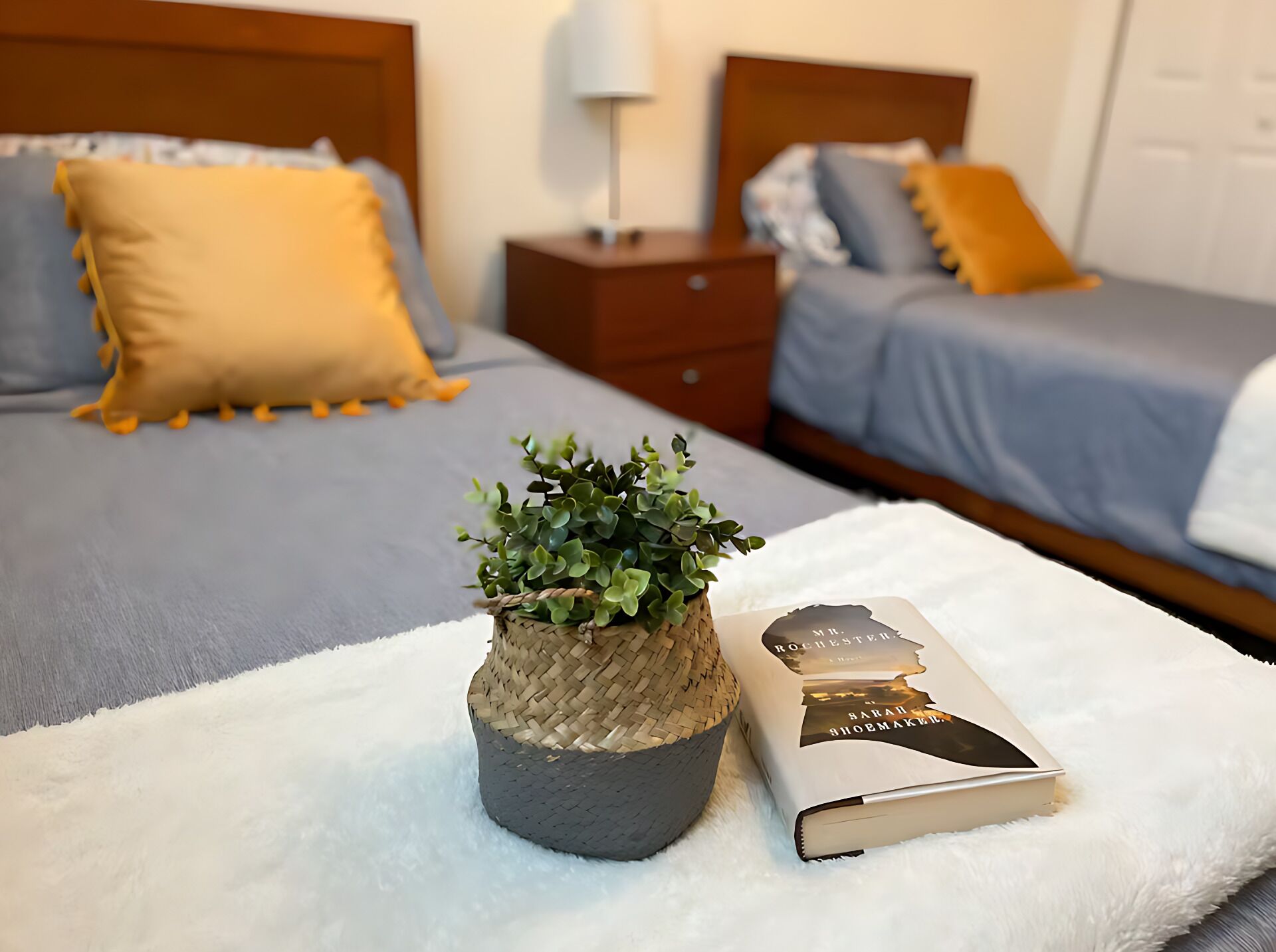 Uma cama bem arrumada com um edredom cinza e azul, um travesseiro amarelo e uma pequena cesta de vime com uma planta verde.