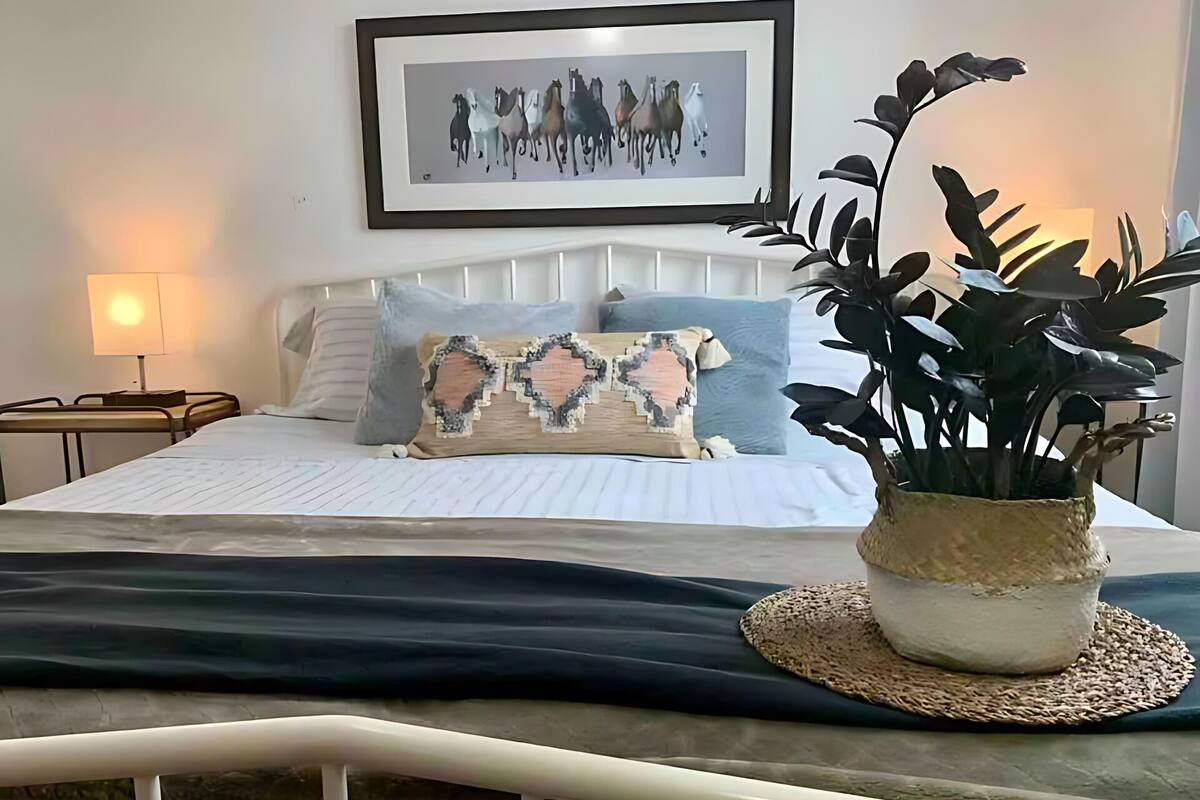 Un lit bien fait, agrémenté d’un coussin décoratif, une table de chevet avec une lampe et une œuvre d’art encadrée au mur.
