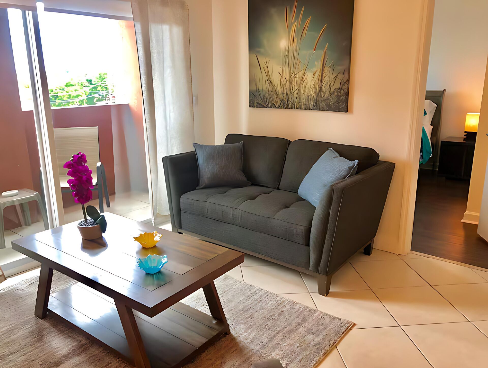 A living room with a grey sofa, a wooden coffee table with decorative items, a painting on the wall, and a view of the outdoors through a glass door.