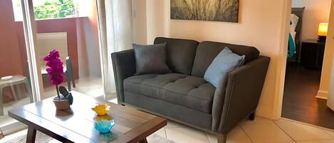 A living room with a grey sofa, a wooden coffee table with decorative items, a painting on the wall, and a view of the outdoors through a glass door.