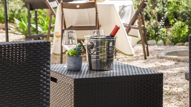 A wicker table with a metal bucket and a small potted plant, set outdoors with a tent and chairs in the background.