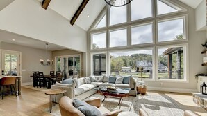 A spacious living room with a large sectional sofa, a glass coffee table, and a view of a house through large windows.