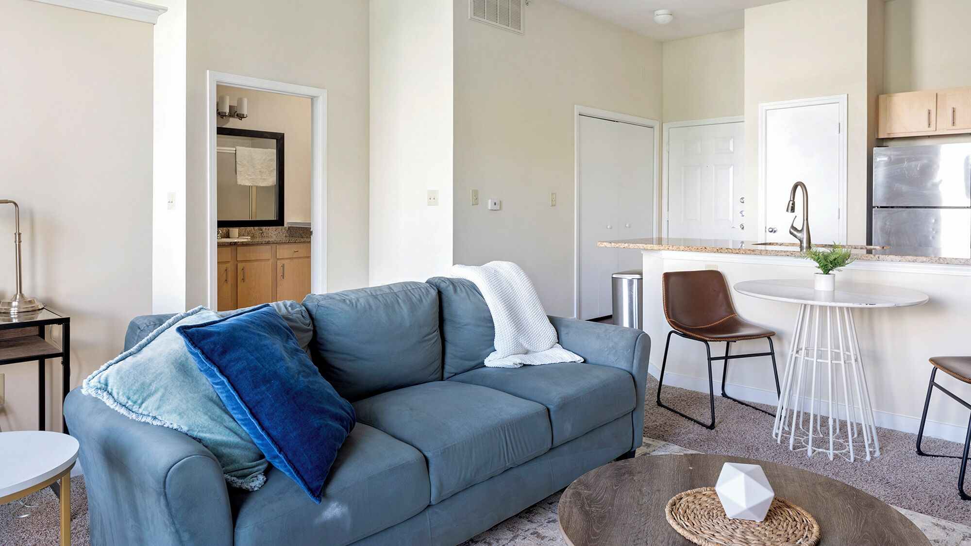 A modern living room with a grey sofa, a coffee table, and a kitchen area with a white countertop and stainless steel appliances.