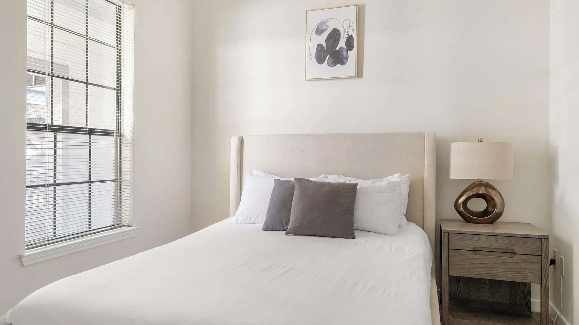 A neatly made bed with white and gray linens, a bedside table with a lamp, and a window with blinds.