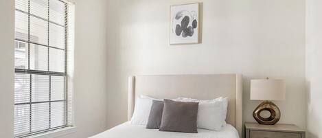 A neatly made bed with white and gray linens, a bedside table with a lamp, and a window with blinds.