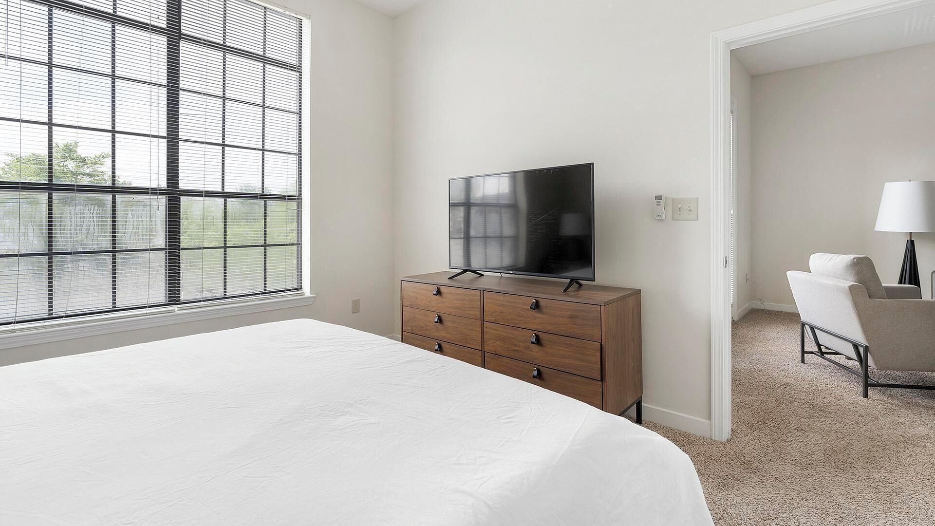 A hotel room with a large bed, a wooden dresser with a flat-screen TV, a chair, and a window with blinds.