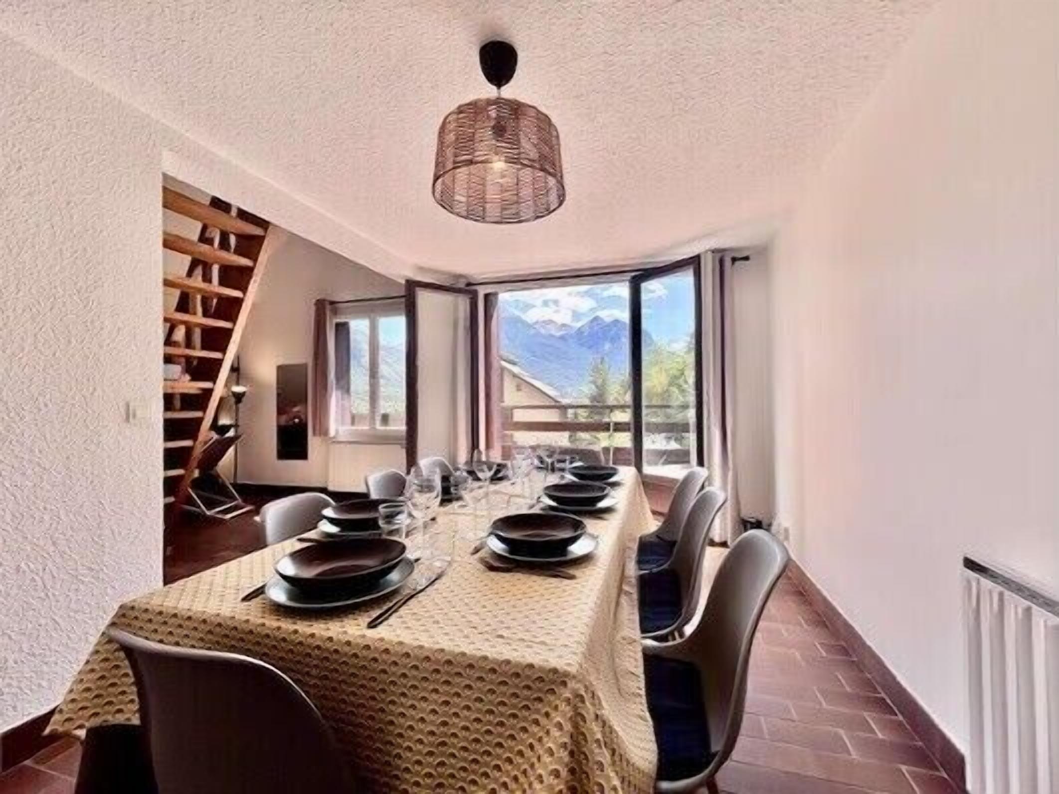 Un espace repas avec une longue table dressée pour quatre personnes, un escalier et une vue sur les montagnes par de grandes fenêtres.