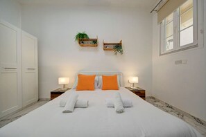 Una habitación de hotel con una cama grande, dos mesitas de noche, una mesita auxiliar, una ventana y una puerta.
