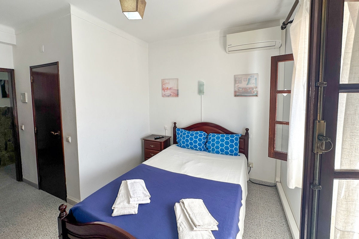 Un dormitorio con una cama, una mesita de noche, aire acondicionado instalado en la pared y una puerta.