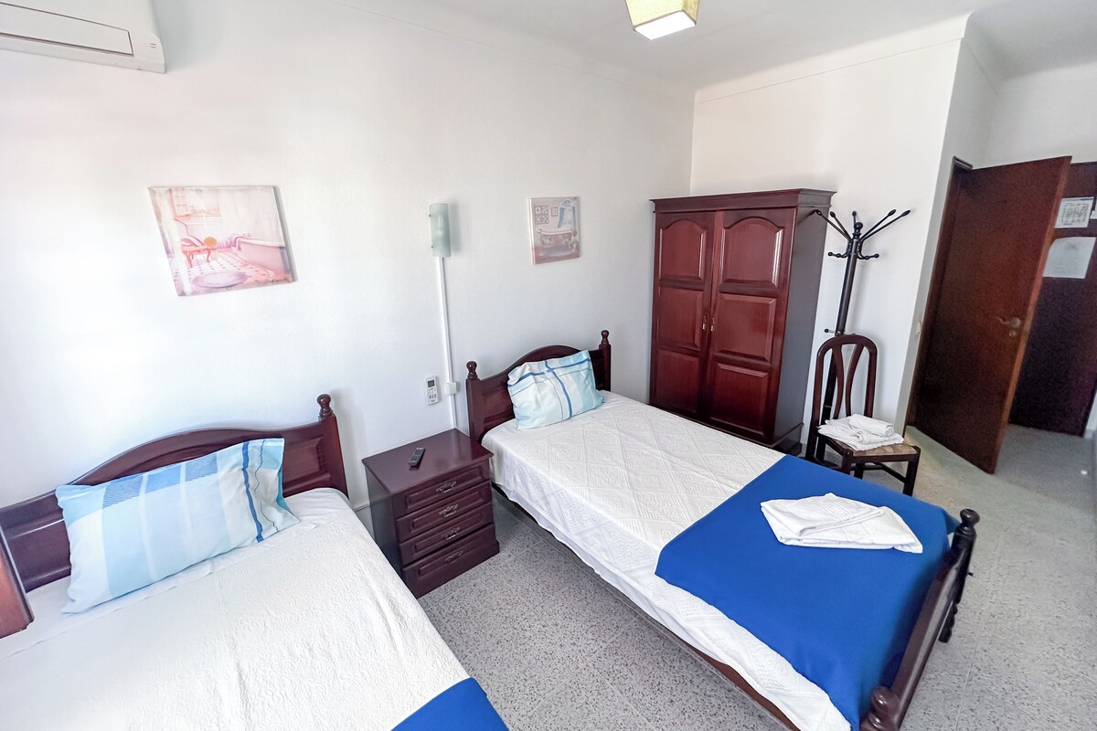 A bedroom with two beds, a wardrobe, a coat rack, and a bedside table with a lamp.