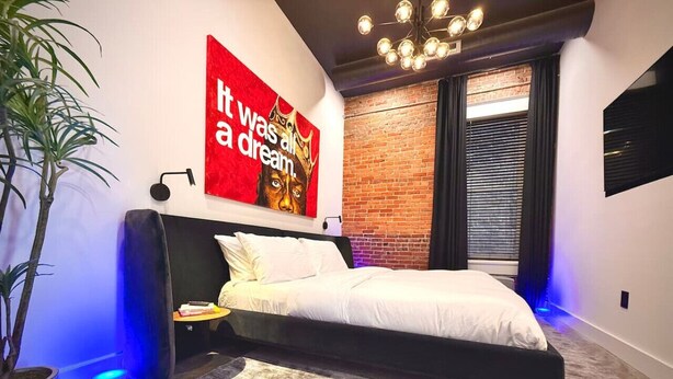 Un dormitorio moderno con una cama, un cartel motivacional rojo, una planta en maceta y una pared de ladrillo.