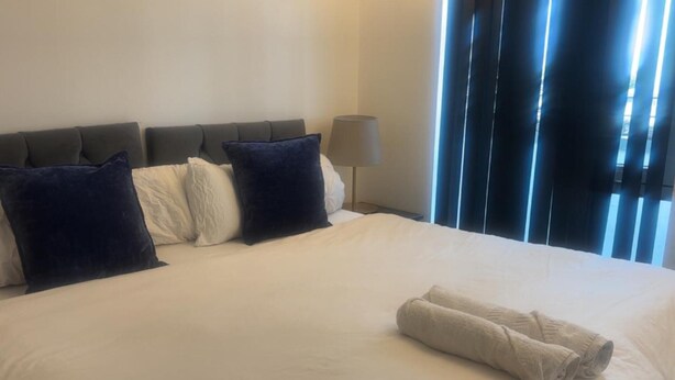 A neatly made bed with a white duvet and two rolled towels, a dark headboard, and a lamp on a bedside table. - London 1 Bed Apartment Close to Barking Station (Barking)