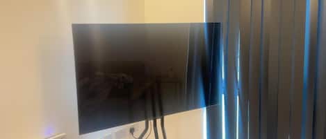 Un téléviseur à écran plat, posé sur un meuble, dans une pièce dotée d’une fenêtre et de stores.