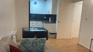 A living room with a sofa, a red pillow, and a patterned throw. A small table with a remote control is on the sofa. The room leads to a kitchen with a clock on the wall and a trash bin. - London 1 Bed Apartment Close to Barking Station (Barking)
