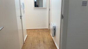 A narrow hallway with wooden flooring, a white door on the left, and a white wall with a small window and a radiator on the right. - London 1 Bed Apartment Close to Barking Station (Barking)