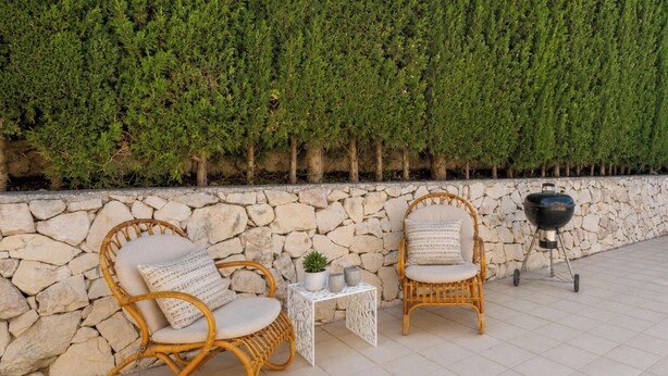 Deux fauteuils en osier avec des coussins, une petite table sur laquelle est posée une plante en pot, et un barbecue adossé à un mur de pierre, le tout agrémenté d’une haie.